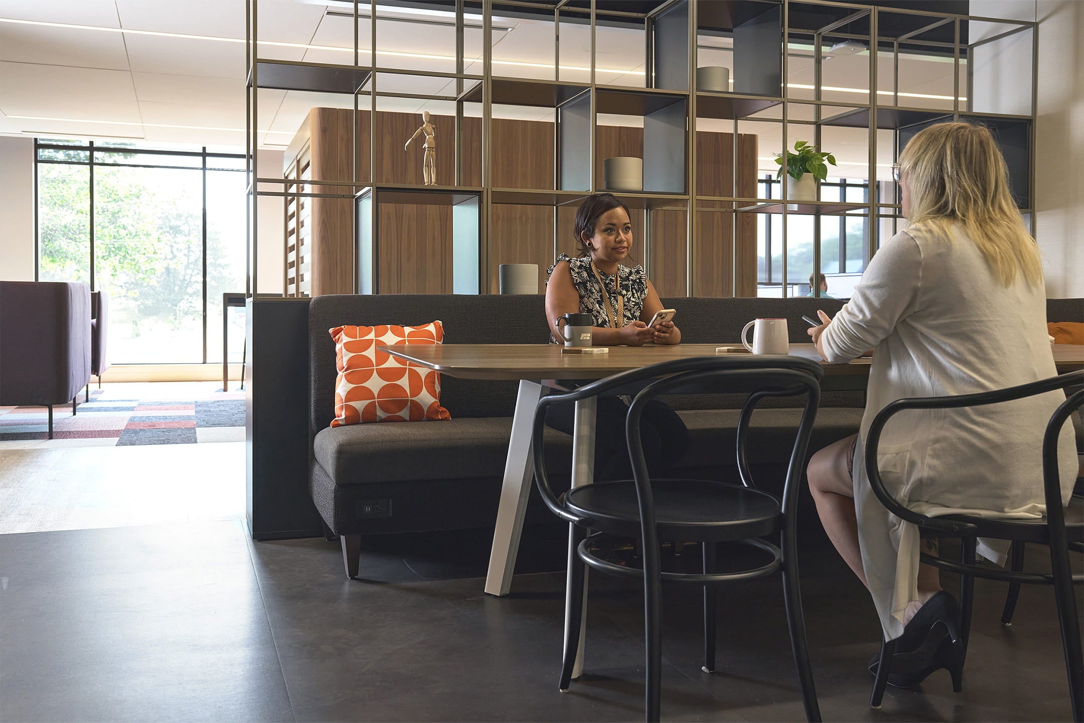 Two women sitting at a table in a modern office setting with large windows and a glass wall.