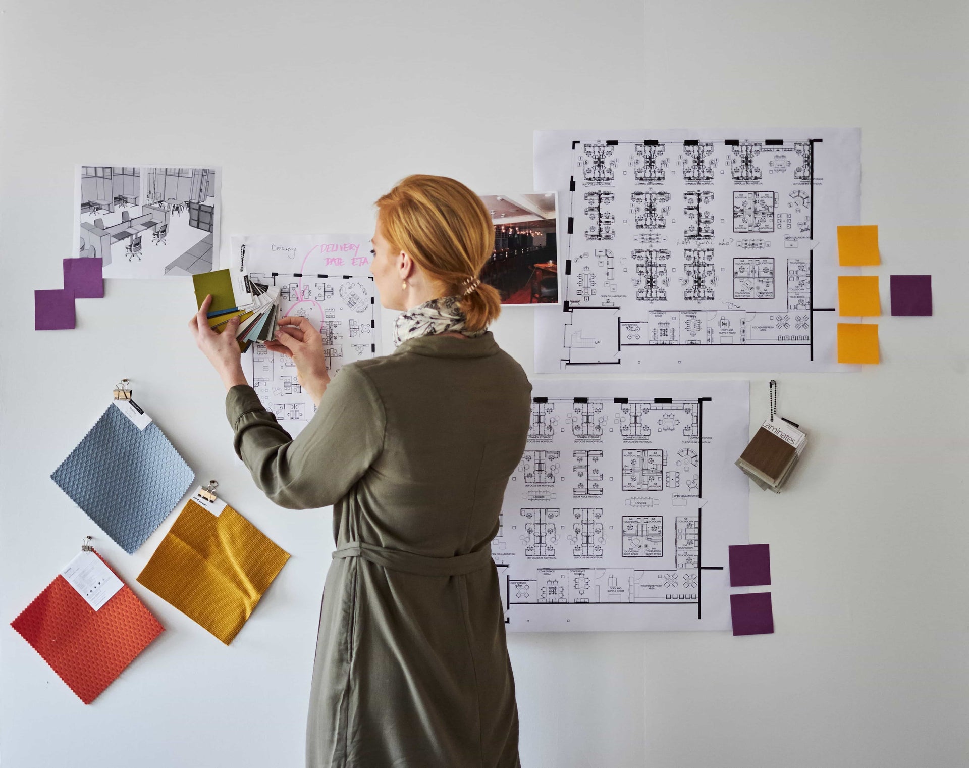 Woman working on a design project with blueprints and color swatches on a white wall.
