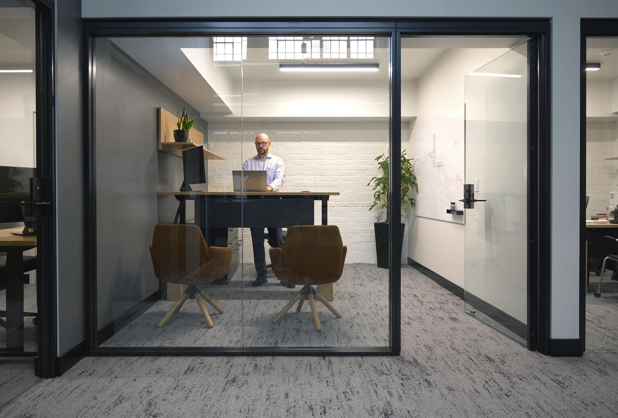 Person working at a desk inside a glass office pod with another person in the background.