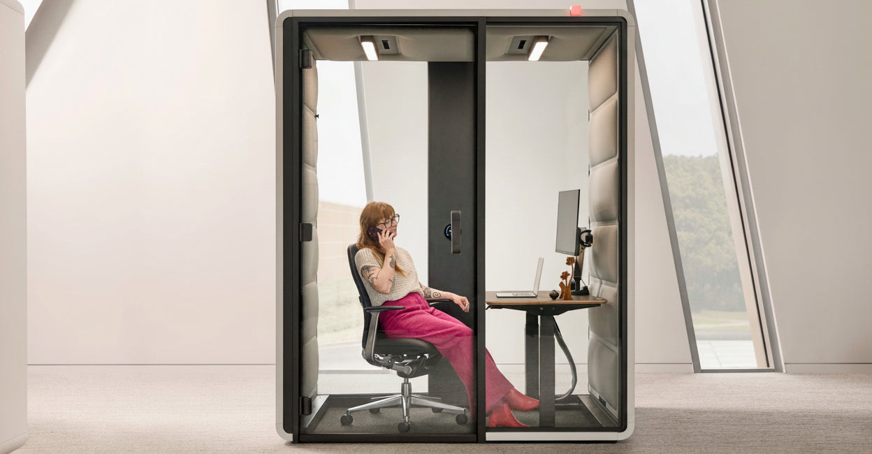 A modern glass office privacy pod with a black frame, shown with a woman sitting inside on a chair at a desk. The pod is set in a minimalist, bright room with large angled windows.
