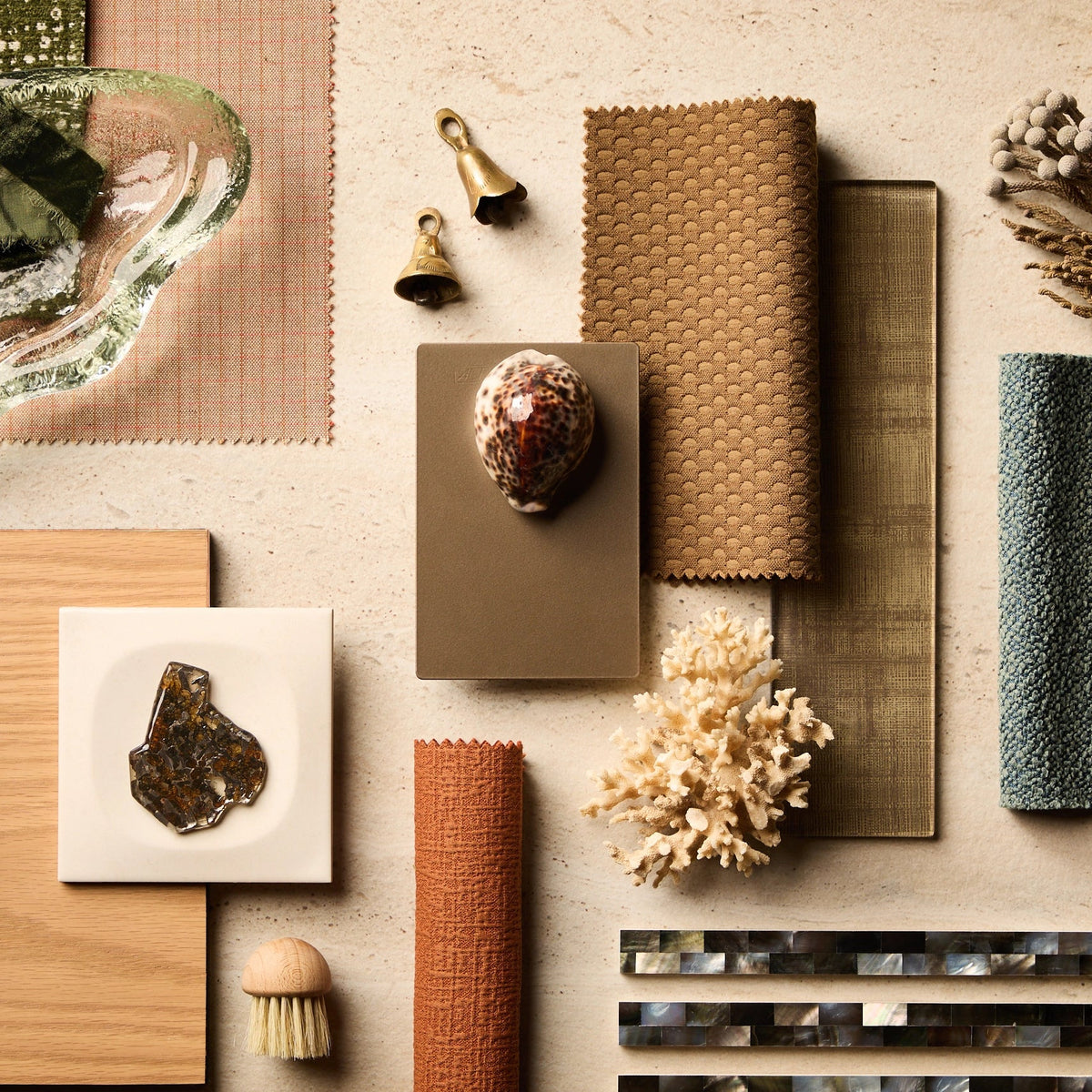 A styled flat-lay of home decor and material samples on a light beige surface, including woven textiles, a glass dish, dried feathers, a shell, a small coral piece, a brush, and tile samples.