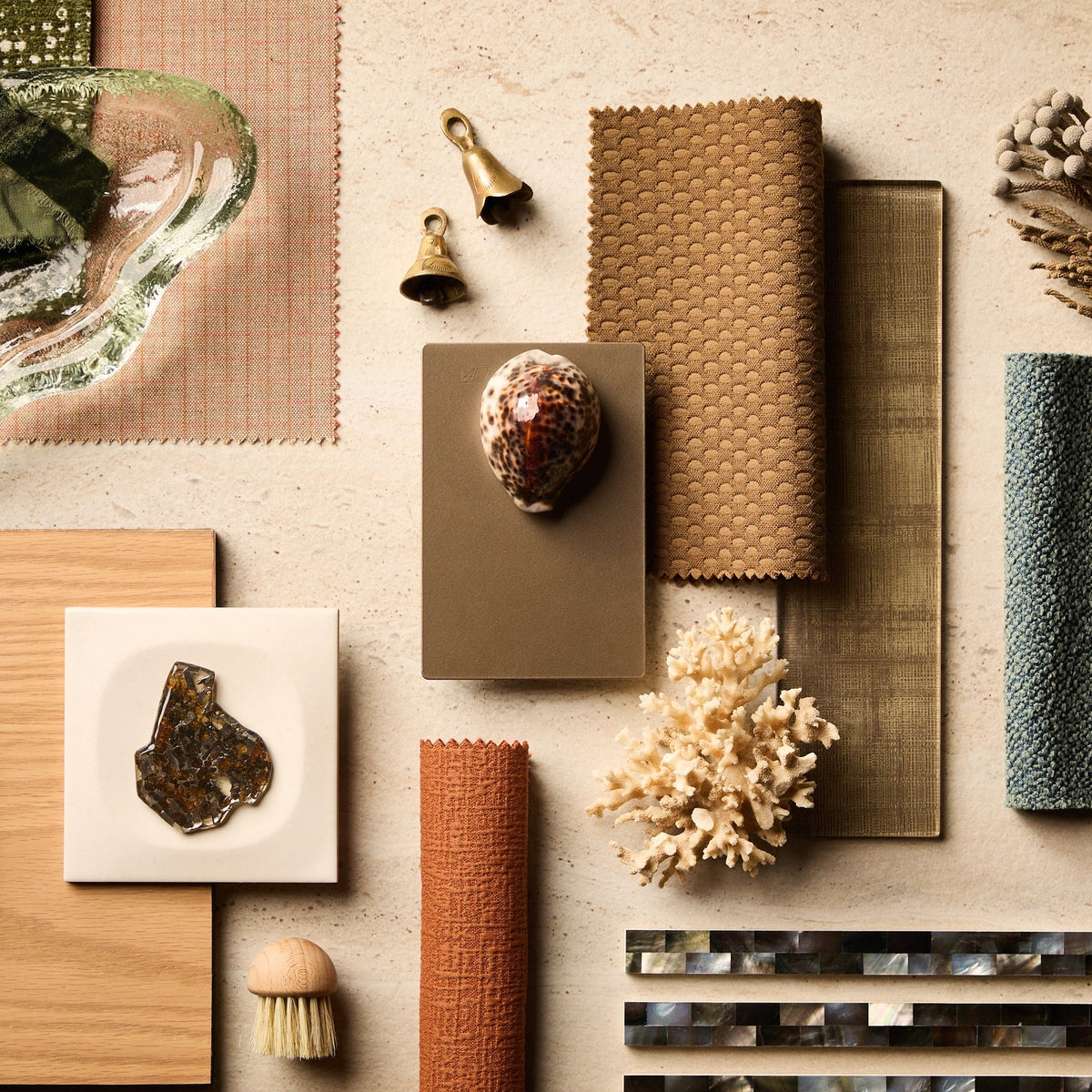 A styled flat-lay of home decor and material samples on a light beige surface, including woven textiles, a glass dish, dried feathers, a shell, a small coral piece, a brush, and tile samples.