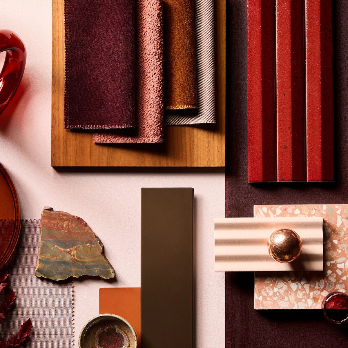 A styled flat-lay arrangement of home decor and materials in warm earthy tones, including stacked fabric or leather swatches on a wooden board, red striped panels, a marbled stone slab, ceramic tiles, dried botanicals, and amber glassware on a split cream and dark brown background.