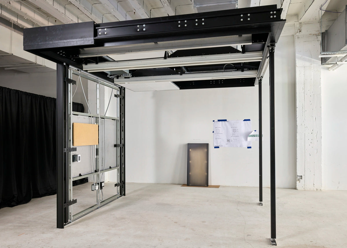 A large black metal gantry crane or industrial frame structure with glass panels, photographed inside a white studio or warehouse space with concrete floor and exposed ceiling beams.