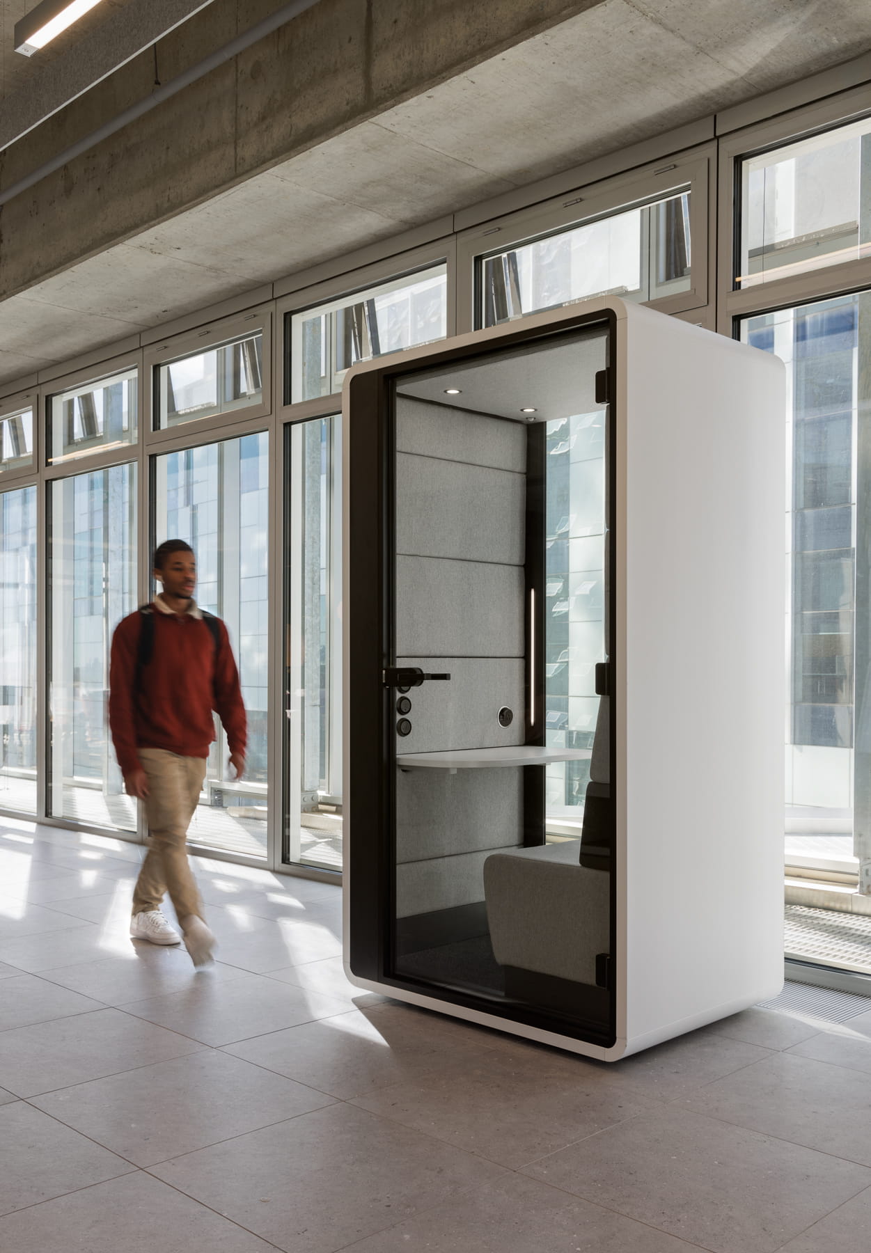 Person walking past a modern white office pod in a bright room with large windows.